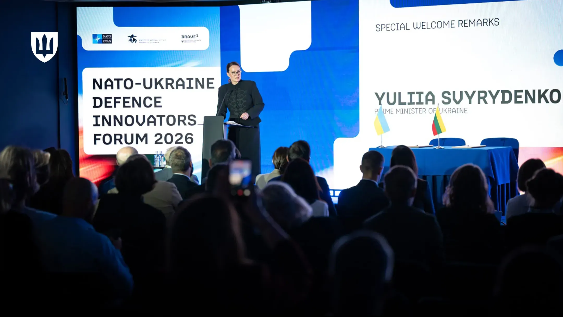 Жінка в темному діловому вбранні виступає за трибуною на сцені конференц-залу перед великою аудиторією слухачів. На екранах позаду неї розміщено англомовні написи про оборонний форум Україна-НАТО 2026 року.