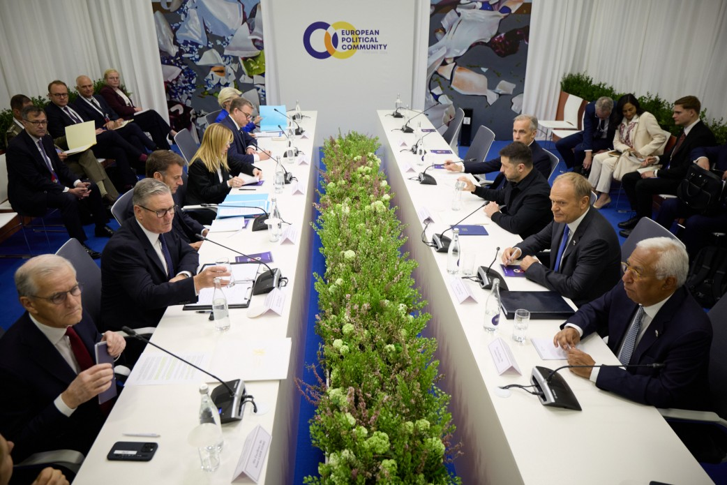 Офіційна зустріч двох делегацій за паралельними довгими столами, між якими висаджено смугу зелених рослин і квітів. На стіні по центру розміщено логотип «European Political Community», а учасники переговорів сидять один навпроти одного з документами, мікрофонами та пляшками води.