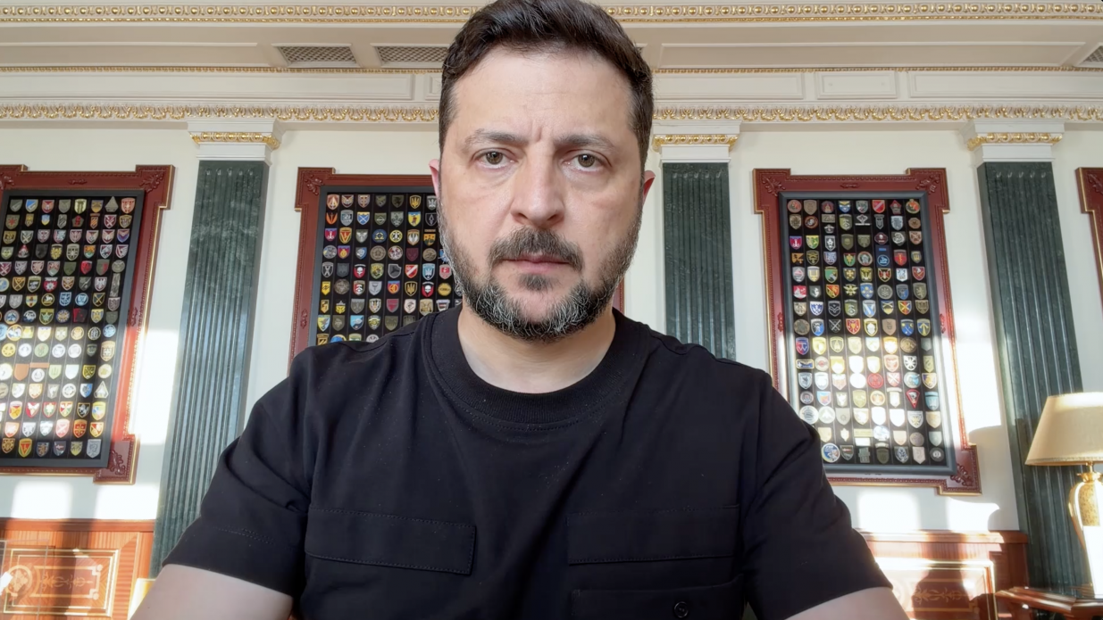 Чоловік із короткою стрижкою та бородою у чорній футболці дивиться прямо в камеру крупним планом. Він перебуває в офіційному кабінеті з класичним інтер'єром, де на задньому плані видно колони та великі настінні стенди з численними військовими шевронами й емблемами.
