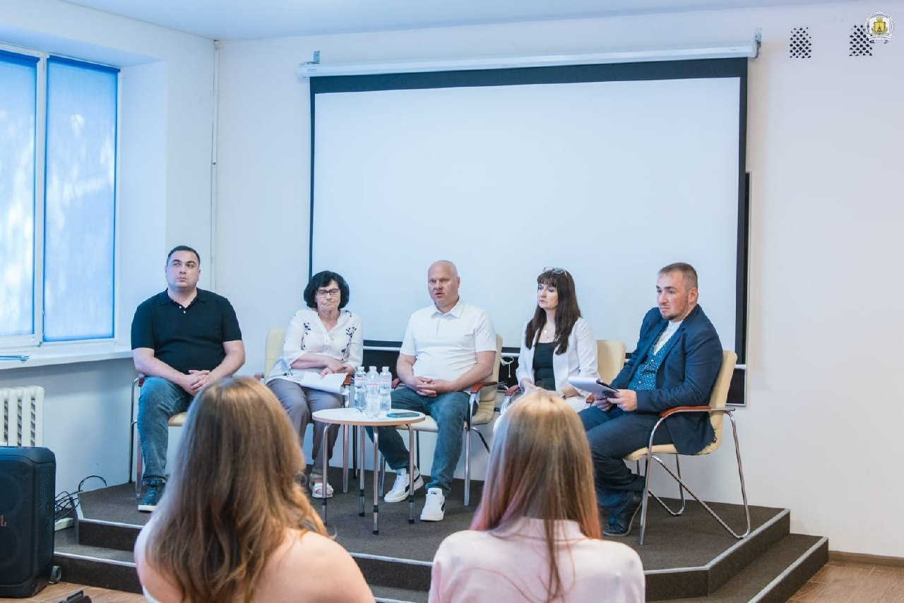 П'ятеро спікерів сидять у кріслах на подіумі перед білим екраном під час офіційної зустрічі. Перед ними стоїть круглий столик із водою, а на передньому плані видно слухачів, які сидять спиною до камери.