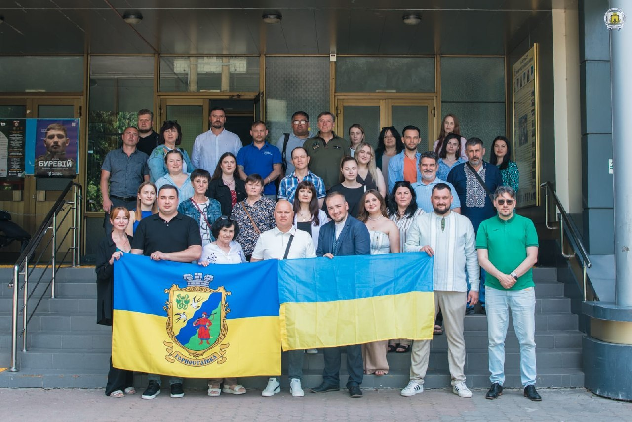 Велика група людей стоїть на сходах біля входу до будівлі для спільного фото. Попереду вони тримають розгорнутий прапор Горностаївки та державний прапор України.