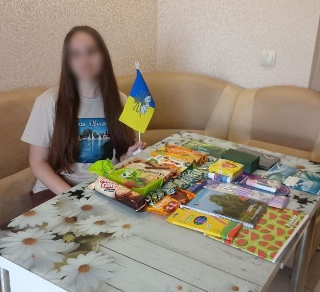 Дівчина з довгим волоссям сидить за кухонним столом із квітковим принтом і тримає маленький прапорець України. На столі перед нею розкладені пакунки солодощів, цукерки, а також шкільне приладдя: олівці, зошити та блокноти.