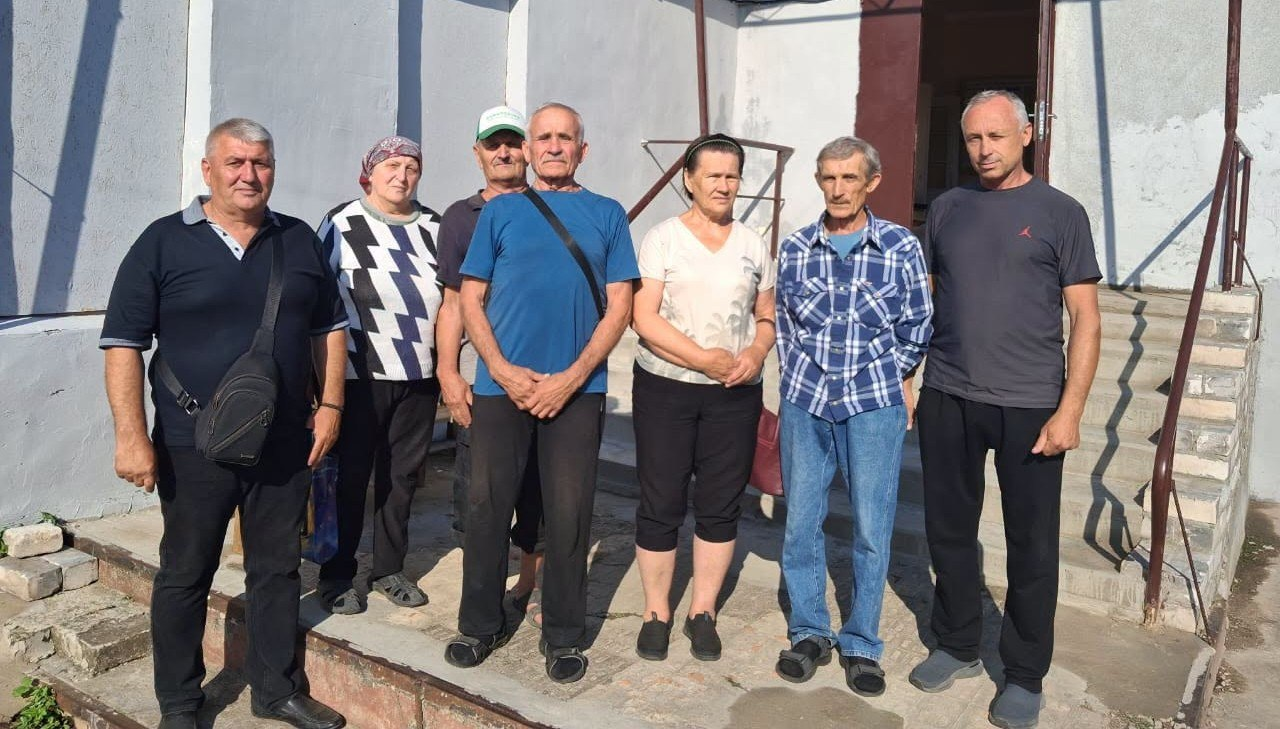 Група з семи дорослих людей (чотирьох чоловіків та трьох жінок) стоїть у ряд на тлі світлої стіни біля входу до будівлі з бетонними сходами. Вони одягнені у повсякденний літній одяг: футболки, сорочки, штани та капрі. На вулиці сонячно, праворуч від людей видно відкриті вхідні двері.