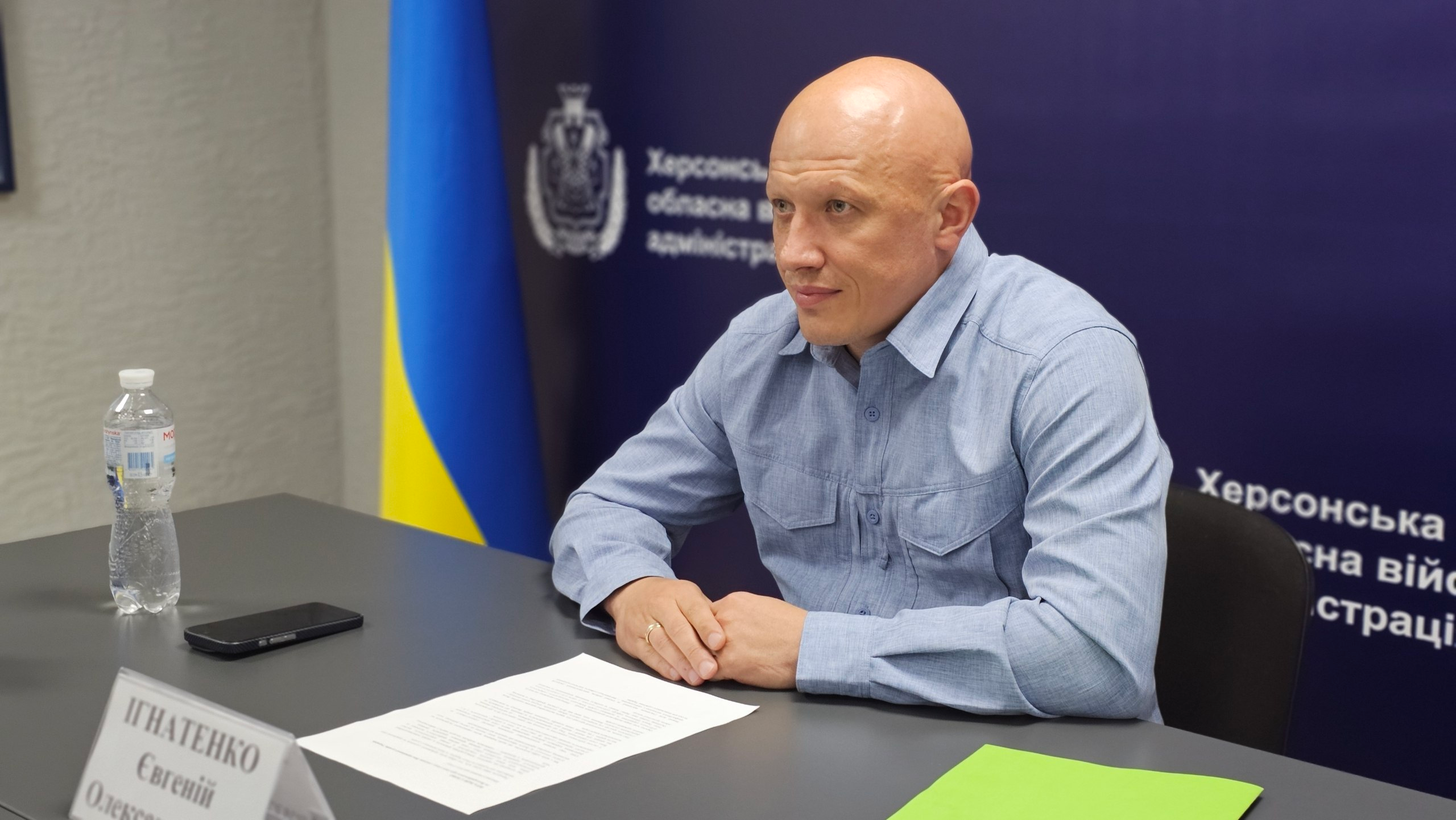 Чоловік у блакитній сорочці сидить за сірим столом і слухає співрозмовника під час офіційної зустрічі. На столі перед ним лежать документи, смартфон, пляшка води та іменна табличка, а на задньому плані розташовані прапор України та темно-синій банер із написами.