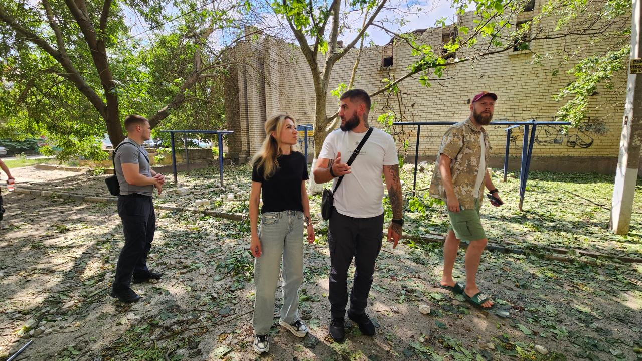 Група людей оглядає подвір'я біля пошкодженої цегляної будівлі після обстрілу. Земля навколо вкрита уламками та зрізаним гіллям дерев.