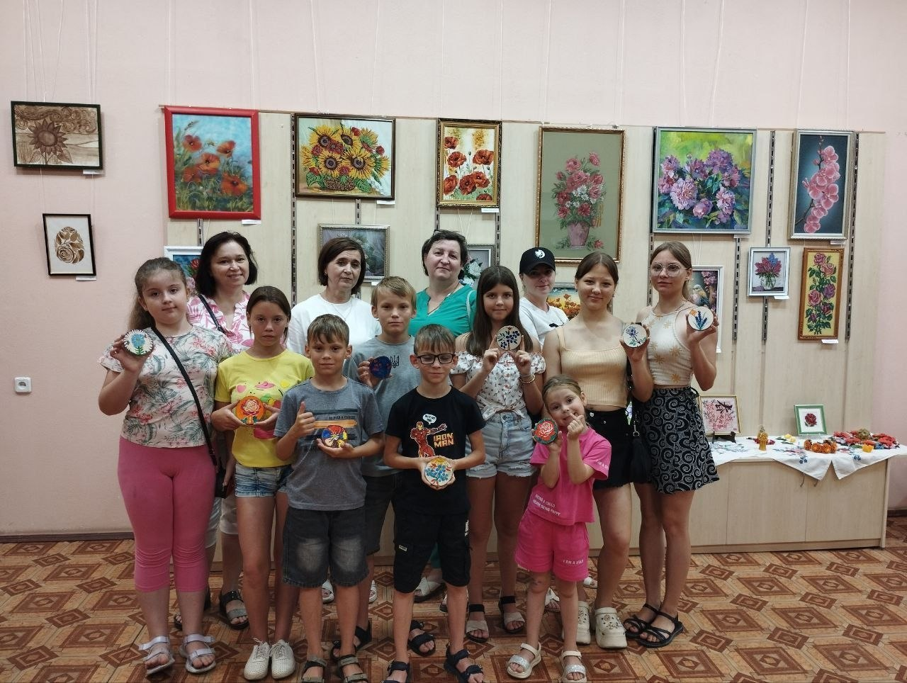Група дітей та дорослих стоїть у виставковій залі на тлі стіни з картинами, тримаючи в руках круглі сувеніри ручної роботи. На задньому плані видно стіл із виробами та різноманітні художні роботи у рамах.