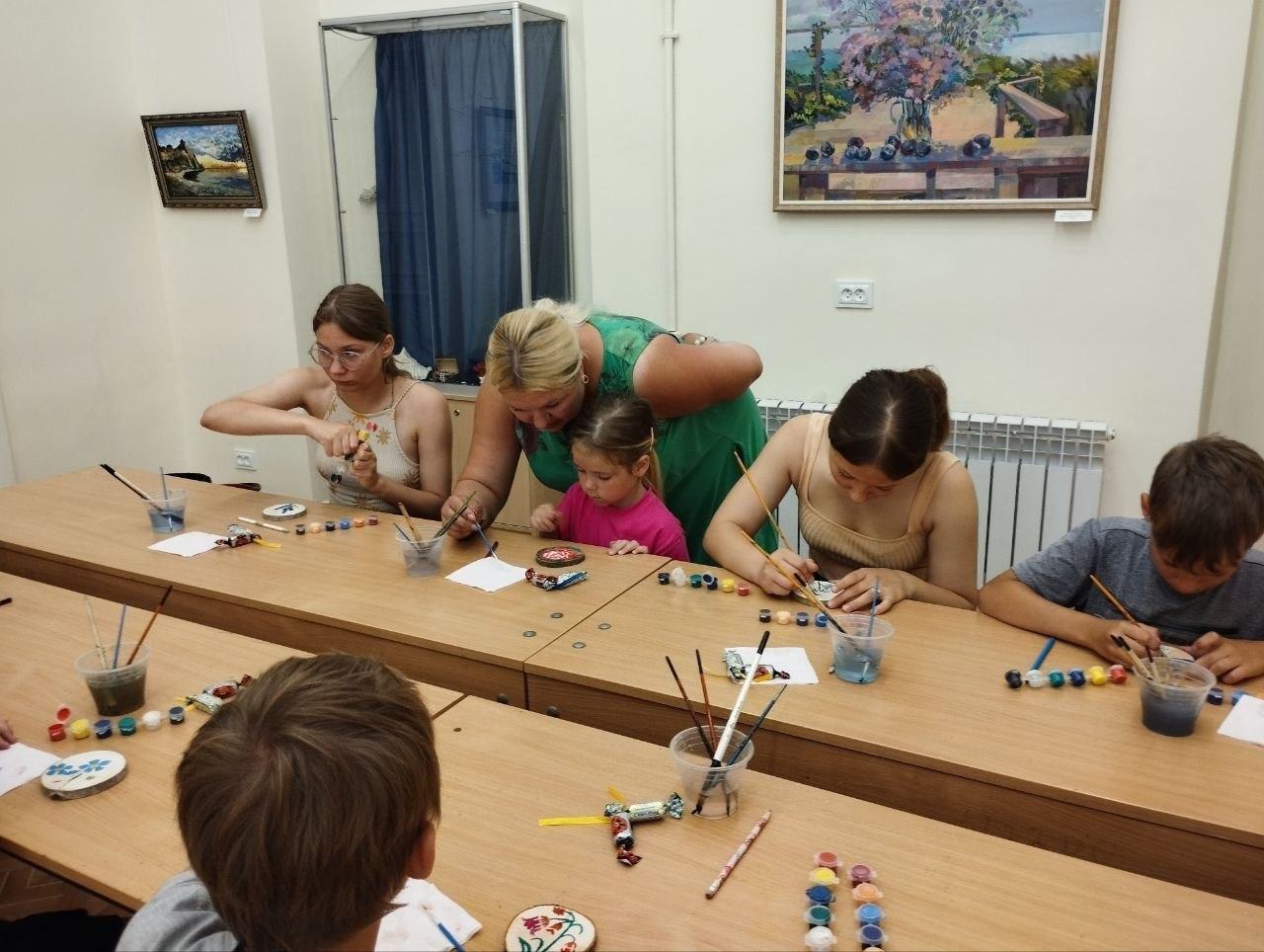 Наставниця в зеленій сукні допомагає дівчинці розмальовувати сувенір під час творчого заняття. Поруч за довгим дерев'яним столом інші діти та підлітки самостійно працюють пензликами та фарбами, а на стіні висить картина у рамі.