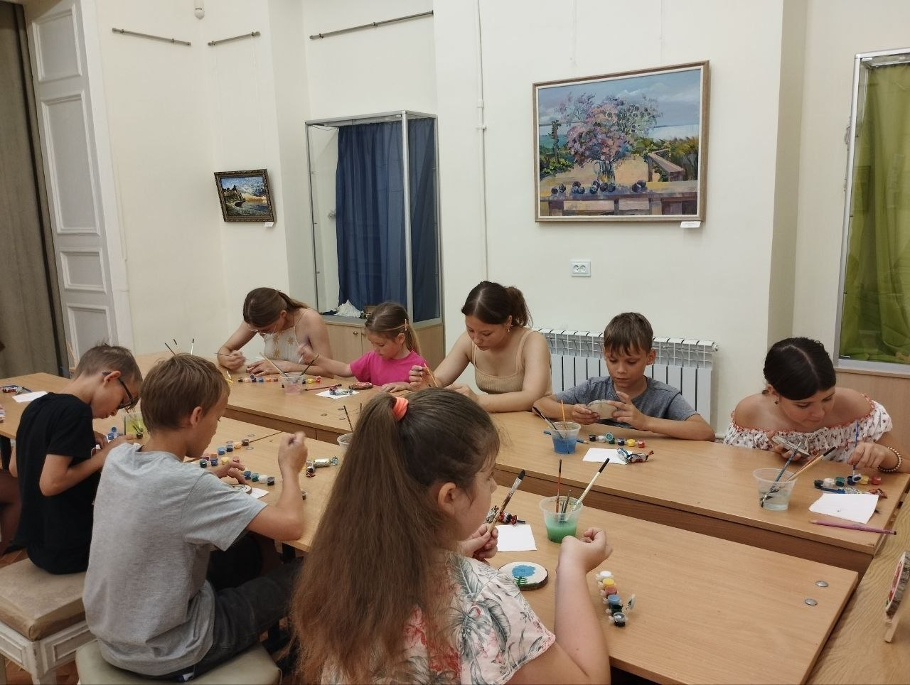Діти та підлітки сидять за довгими дерев'яними столами у світлій кімнаті під час творчого майстер-класу. Вони зосереджено розмальовують маленькі сувеніри фарбами та пензликами, перед кожним стоять баночки з водою та палітри. На стінах розвішані картини в рамах.