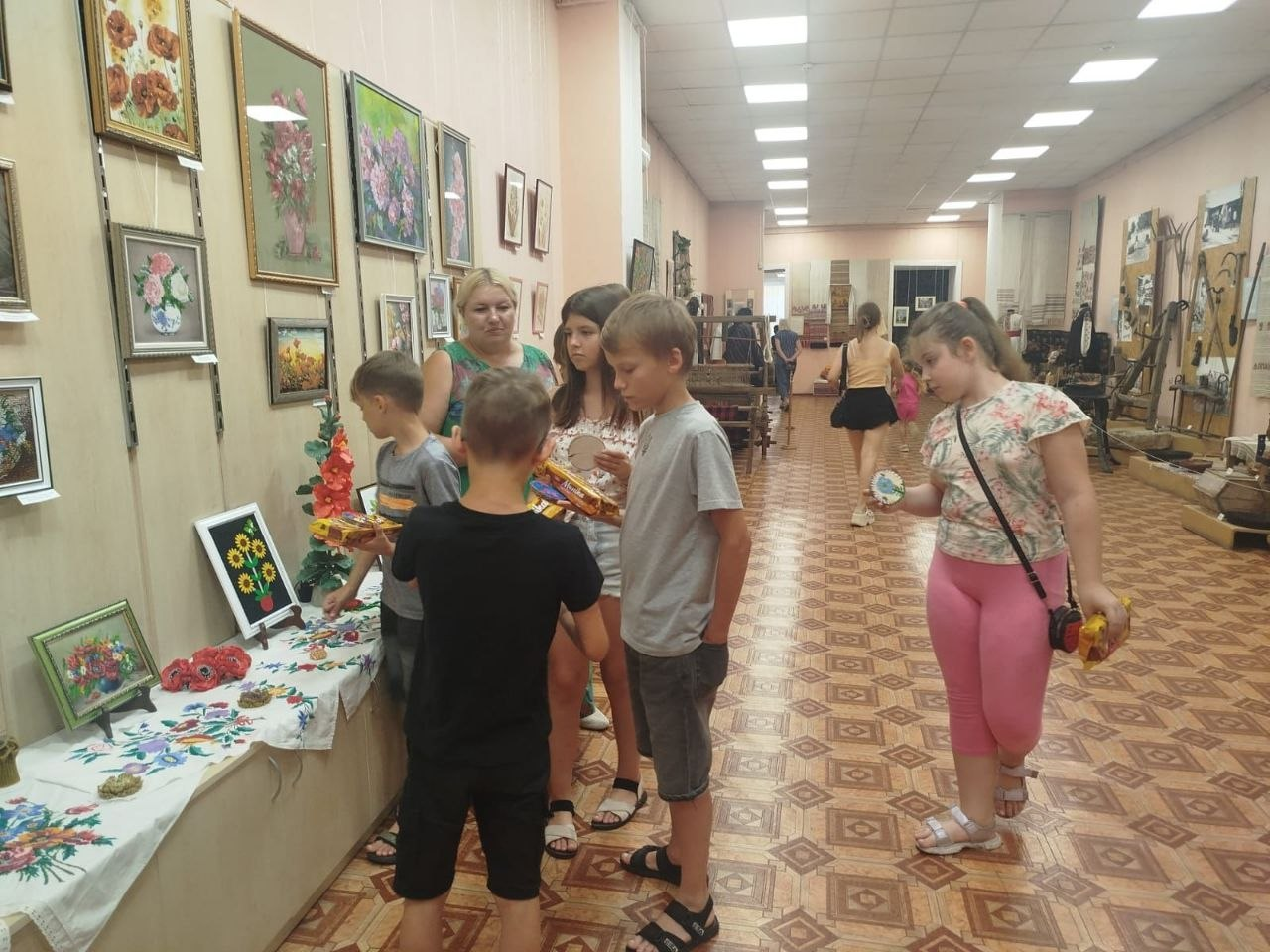Група дітей та наставниця розглядають виставку у довгому музейному залі з візерунчастим лінолеумом. На лівій стіні розвішані картини, а під ними на столі, вкритому вишиваною скатертиною, розкладені сувеніри та вироби ручної роботи.