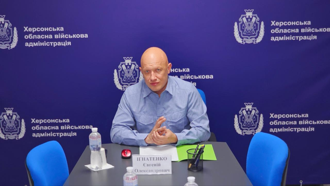 Чоловік у блакитній сорочці сидить за столом з документами та іменною табличкою під час виступу на тлі темно-синього банера з офіційною символікою.
