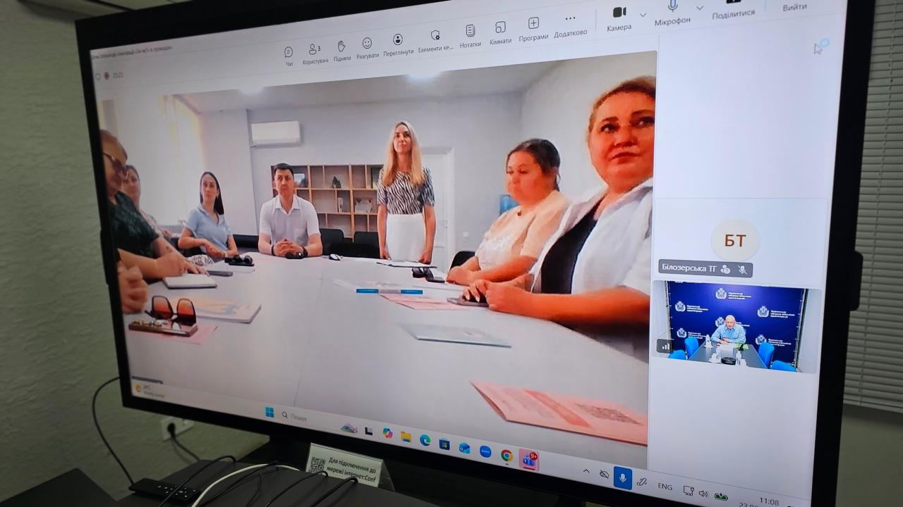 Великий монітор, на якому транслюється онлайн-зустріч в Microsoft Teams. У головному вікні видно групу учасників, які сидять за робочим столом у кабінеті, а в бічній панелі відображаються інші підключені користувачі конференції.