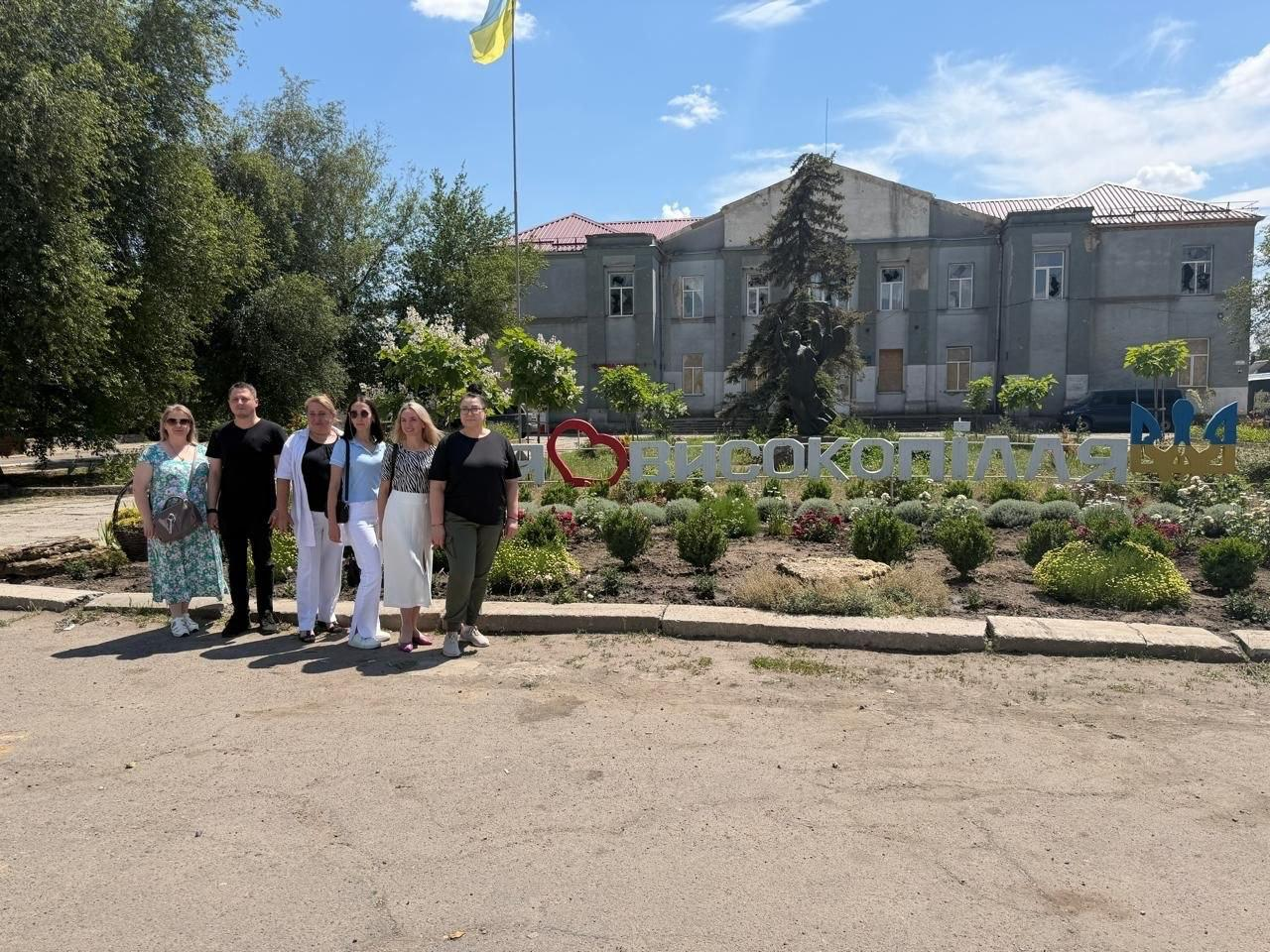 Група людей стоїть на вулиці для спільного фото на тлі великого напису «Високопілля» з червоним серцем. Позаду них облаштована клумба з декоративними кущами, височіє будівля з прапором України на флагштоці, а навколо ростуть зелені дерева.