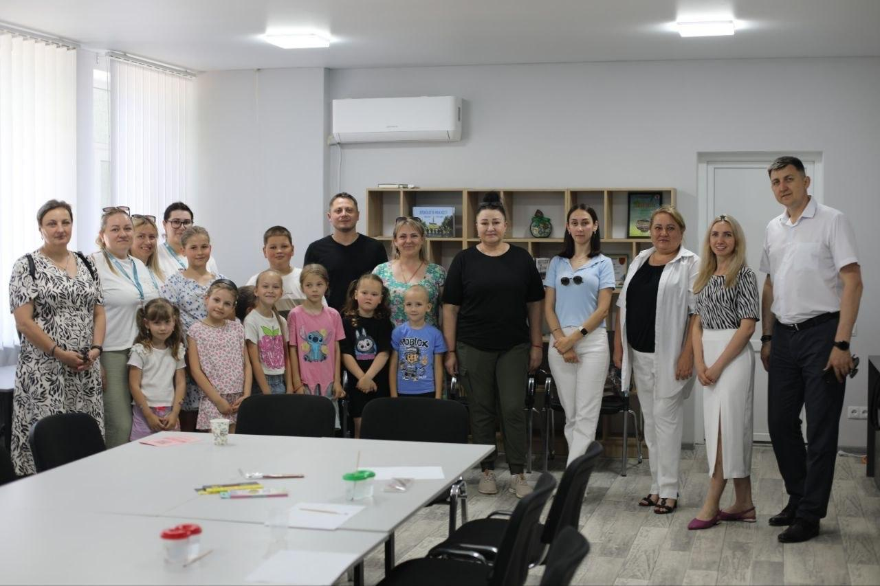 Велика група дітей та дорослих стоїть разом у світлому кабінеті для спільного фото. На передньому плані видно великий робочий стіл зі стільцями та приладдям для малювання, а на задньому плані — відкритий дерев'яний стелаж із книгами та сувенірами.