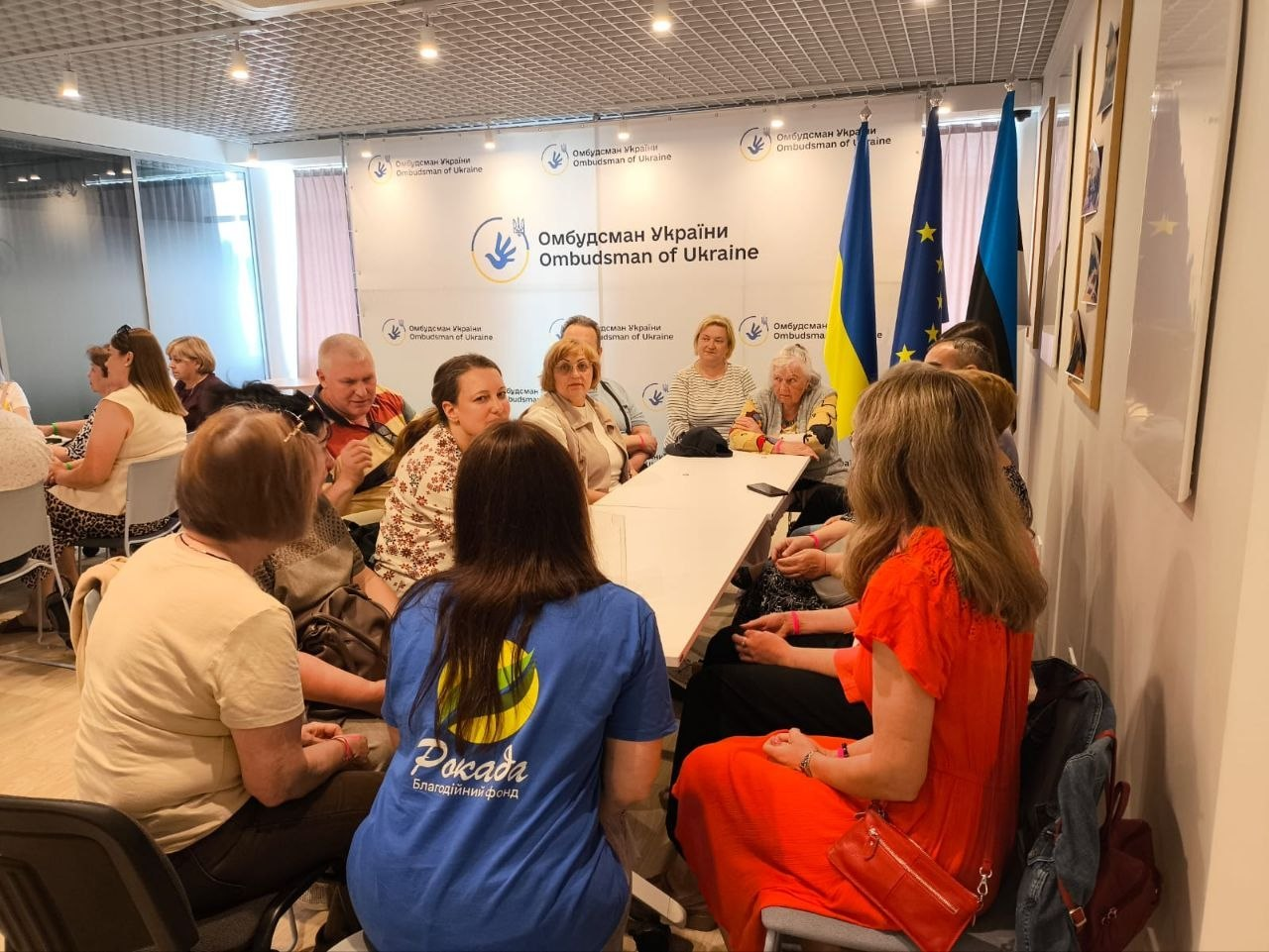Група людей сидить за довгим білим столом під час зустрічі на тлі банера «Омбудсман України / Ombudsman of Ukraine». На передньому плані видно жінку у синій футболці з логотипом благодійного фонду «Рокада», а праворуч стоять прапори України та Європейського Союзу.