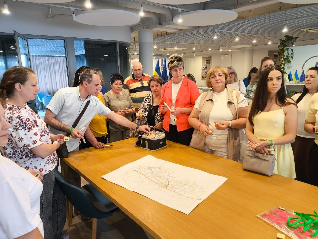Група людей зібралася навколо великого дерев'яного столу під час заходу або тренінгу в сучасному офісному приміщенні. На столі розкладено великий аркуш паперу зі схематичним малюнком чи картою, а один із чоловіків тягнеться рукою до невеликої коробки по центру.