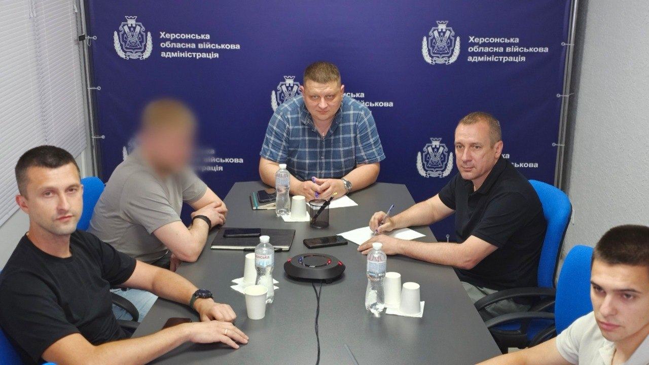 П'ятеро чоловіків сидять за великим сірим столом під час робочої наради. На столі перед ними розкладені папери, ручки, пляшки з водою та паперові стаканчики, а по центру встановлено круглий пристрій для гучного зв'язку. Позаду учасників розташовано темно-синій банер із гербом та офіційними написами.