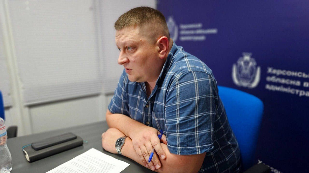Чоловік у синій сорочці в клітинку сидить у кріслі за столом, склавши руки перед собою та тримаючи ручку. На столі перед ним лежать папери та блокнот, а позаду нього видно синій банер з офіційним логотипом та текстом.