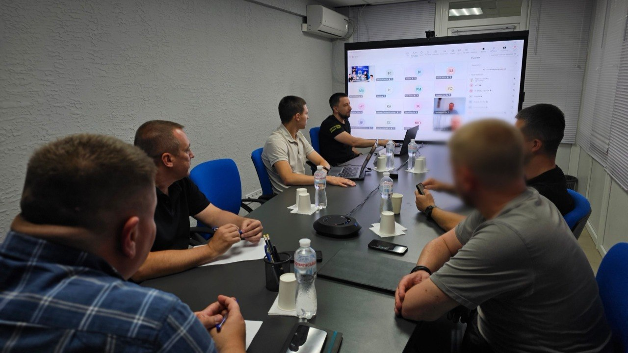 Група чоловіків сидить за довгим столом у робочому кабінеті під час онлайн-наради. Вони дивляться на великий настінний екран, де відображаються вікна учасників відеоконференції. На столі перед ними розкладені ноутбуки, папери, пляшки з водою та паперові склянки.