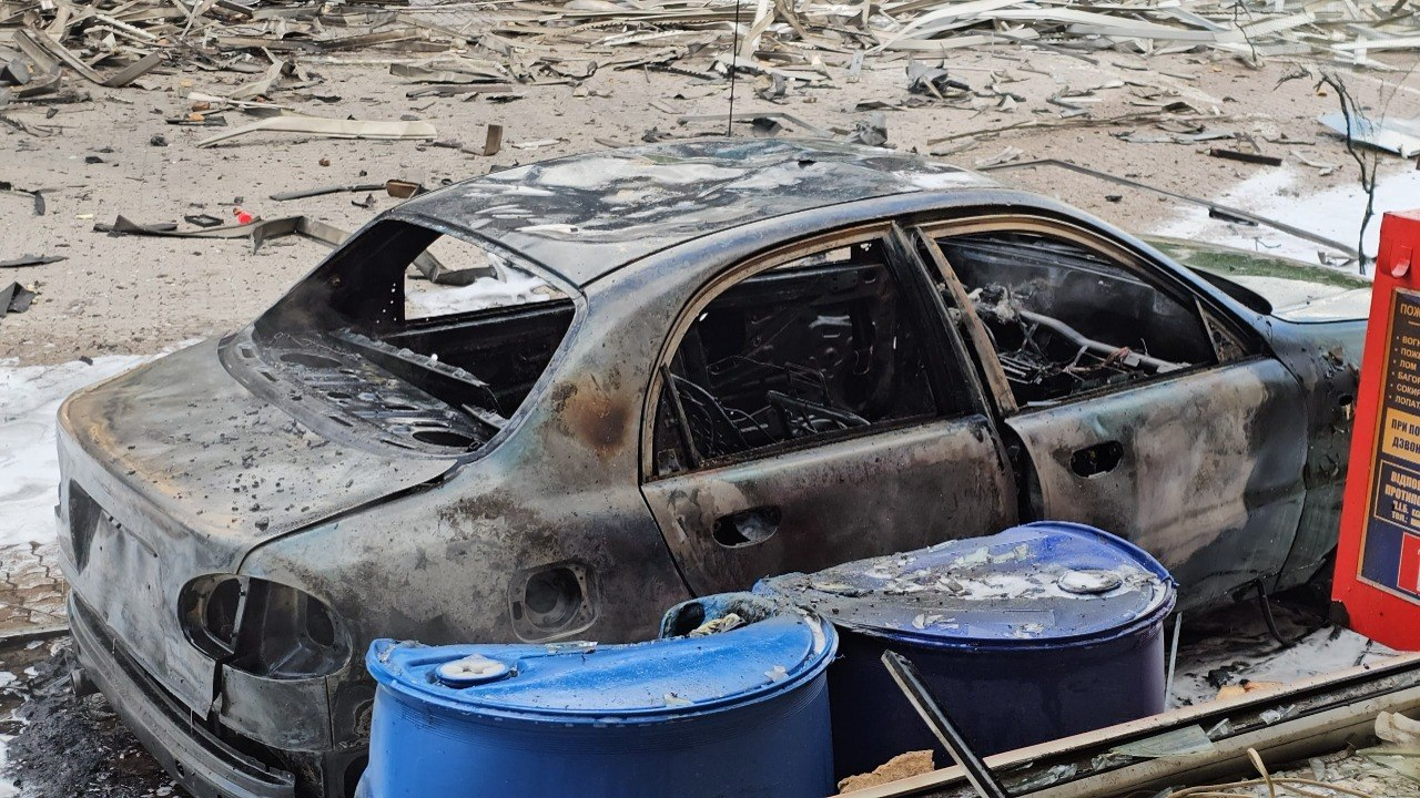 Повністю згорілий легковий автомобіль із вибитим склом та знищеним салоном після обстрілу. На передньому плані перед машиною стоять дві великі сині пластикові бочки, а земля навколо вкрита попелом та численними дрібними уламками.