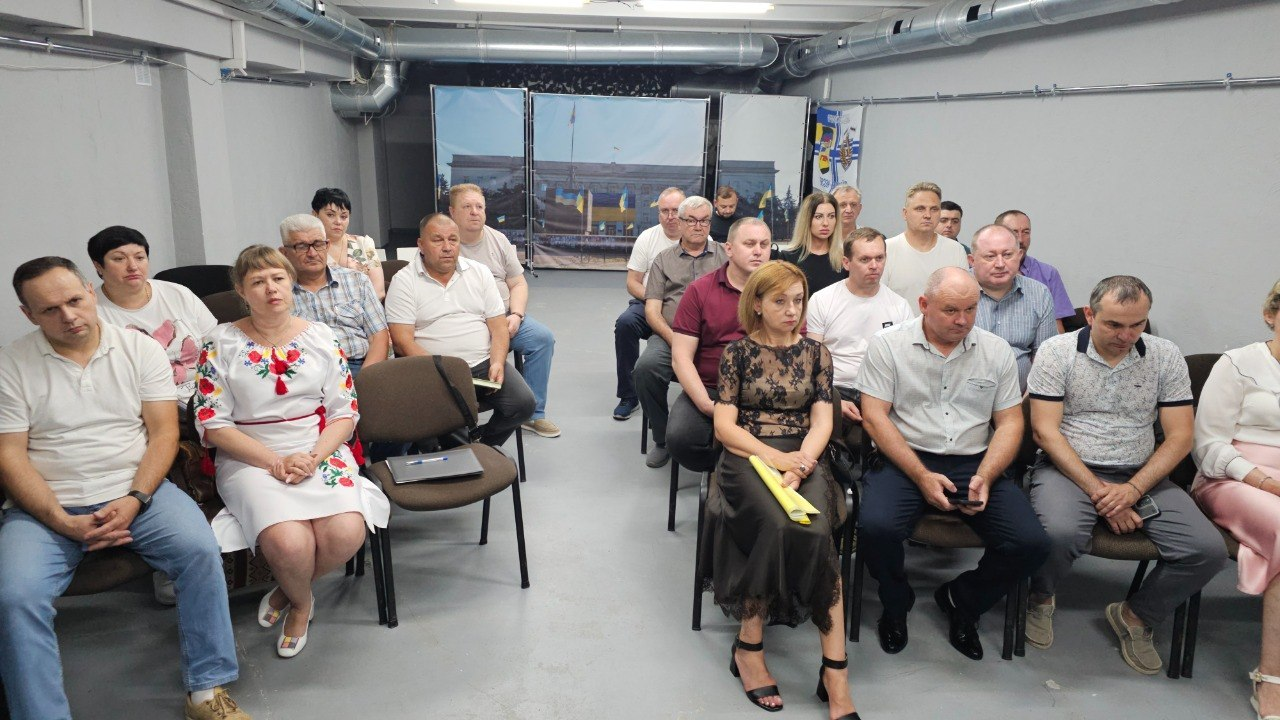 Група людей сидить на стільцях під час заходу в цивільному приміщенні з сірими стінами. Серед присутніх чоловіки та жінки різного віку, а на задньому плані встановлено великий банер із зображенням офіційної адміністративної будівлі з державним прапором України.