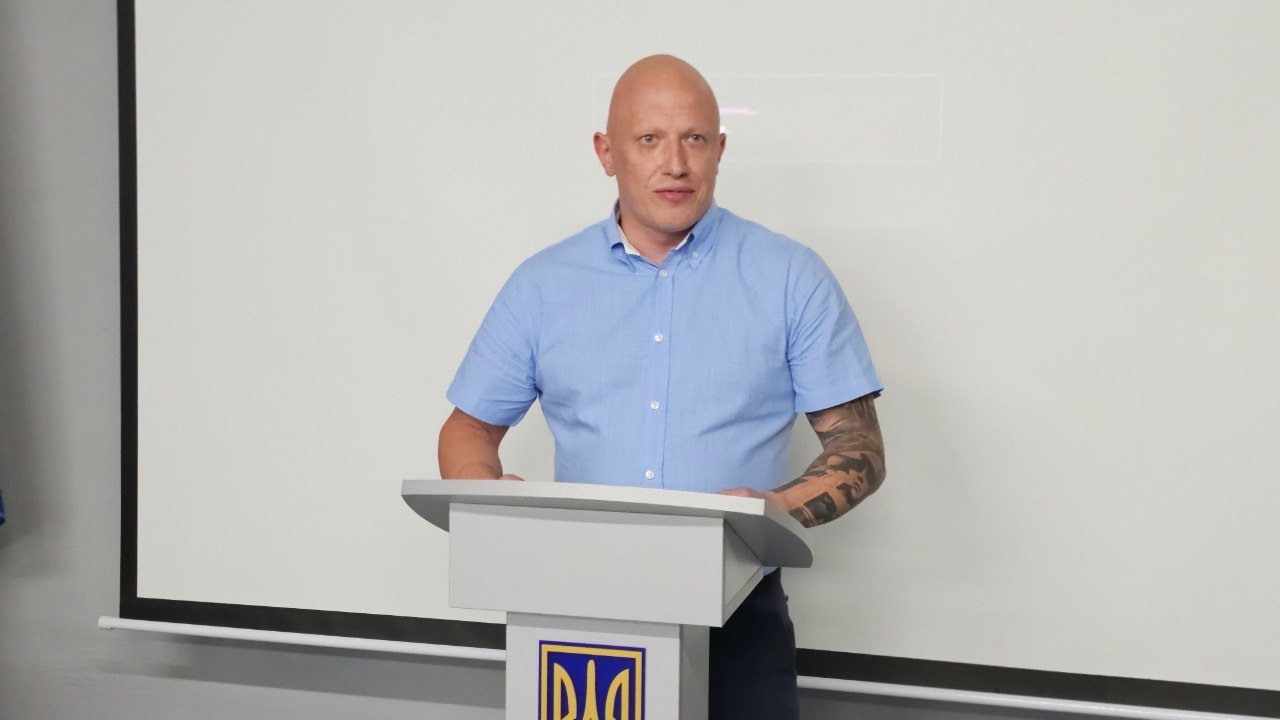 Чоловік виступає за трибуною з державним гербом України на тлі великого білого екрана.