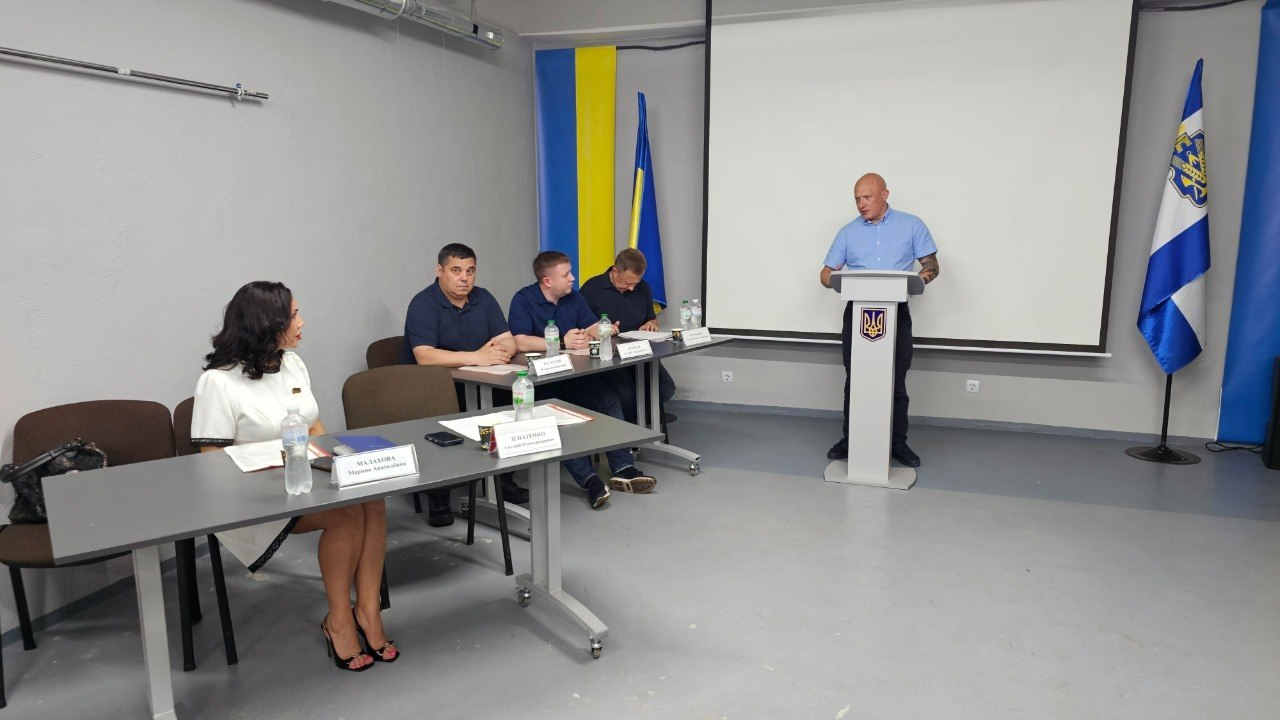 Чоловік виступає за трибуною з державним гербом України перед президією та учасниками заходу в цивільному приміщенні. За столами сидять чоловіки та жінка, поруч встановлені державний прапор України, прапор Херсонської області та великий білий екран.