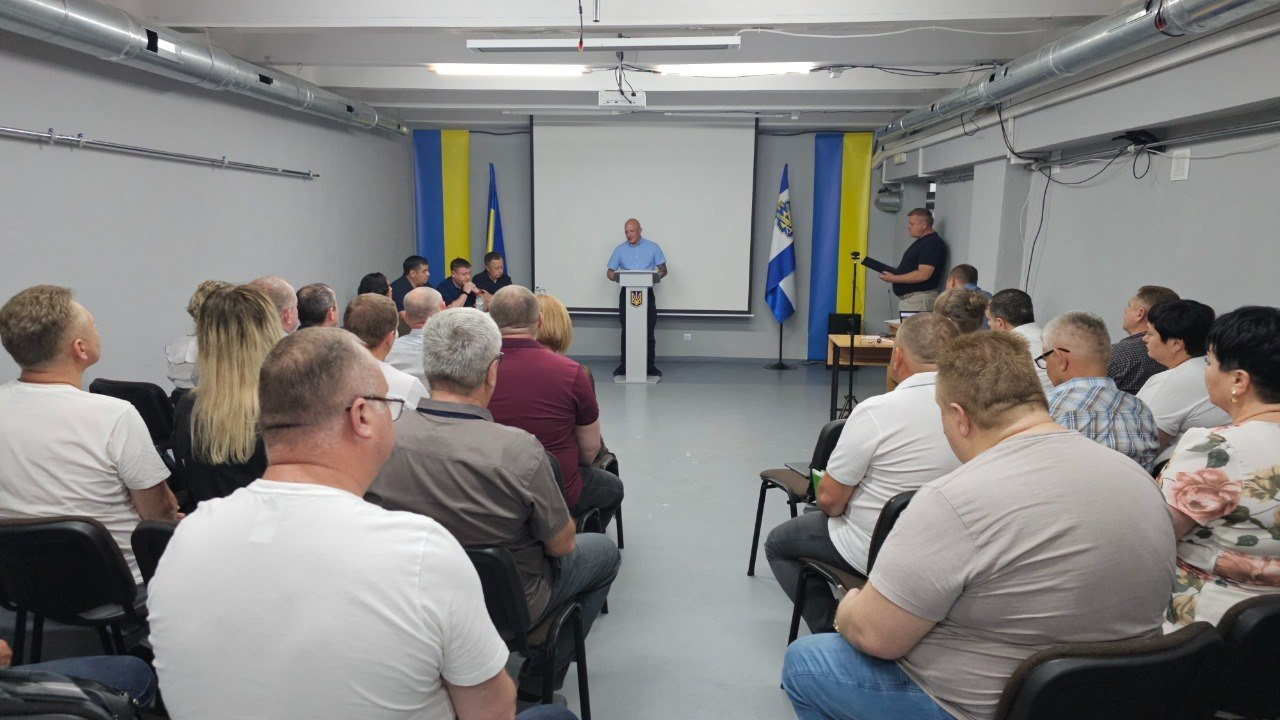 Група людей сидить на стільцях під час заходу в цивільному приміщенні з сірими стінами та слухає чоловіка, який виступає за трибуною з державним гербом України. На задньому плані перед президією встановлені державні прапори України, прапор Херсонської області та великий білий екран.