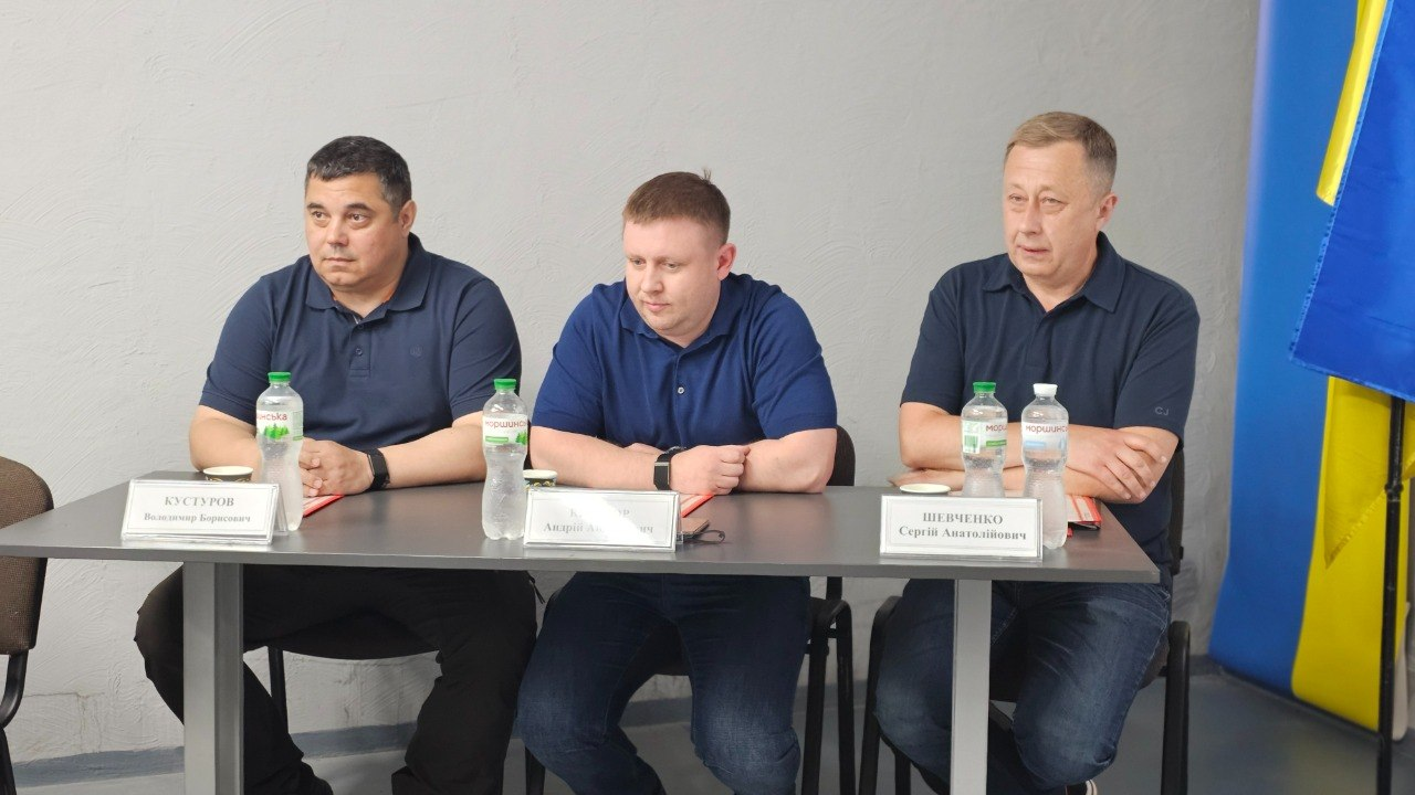 Троє чоловіків сидять за столом під час заходу в цивільному приміщенні з сірими стінами. Перед ними на столі стоять таблички з іменами та пляшки з водою, а праворуч видно частину державного прапора України.