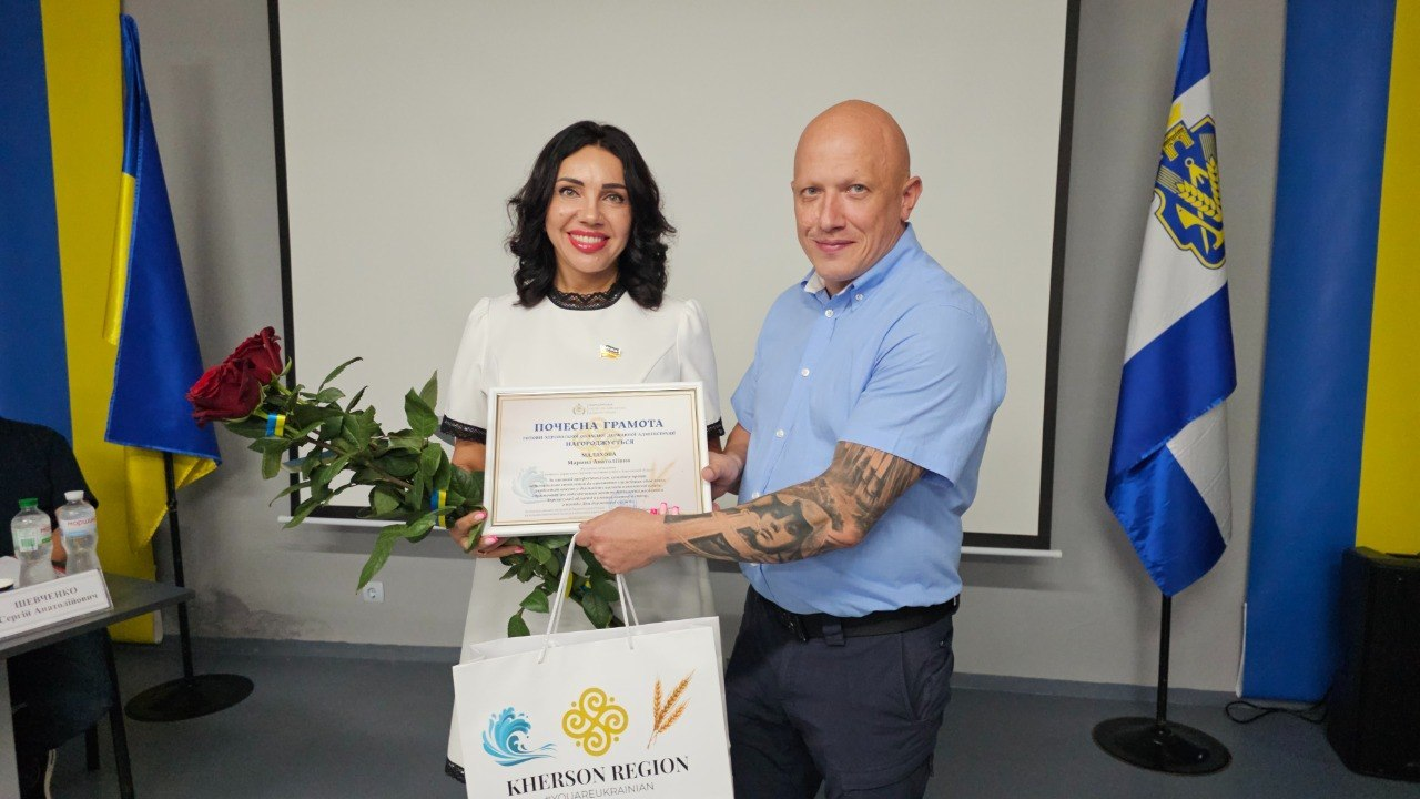 Чоловік та жінка у світлій сукні стоять поруч у приміщенні з сірими стінами та тримають спільну почесну грамоту. Жінка також тримає букет червоних троянд і фірмовий паперовий пакет, а на задньому плані видно великий білий екран, державний прапор України та прапор Херсонської області.