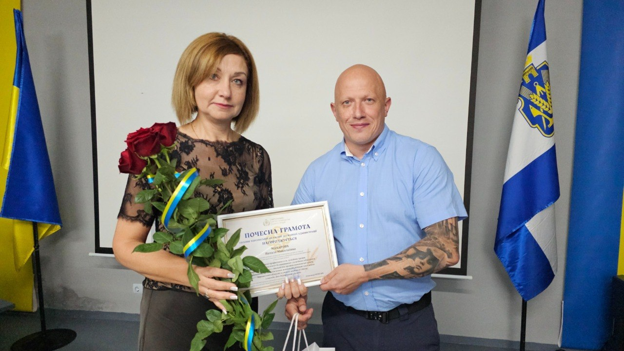Чоловік та жінка в темній блузі стоять поруч у приміщенні з сірими стінами та тримають спільну почесну грамоту. Жінка також тримає букет червоних троянд, а на задньому плані видно великий білий екран, державний прапор України та прапор Херсонської області.