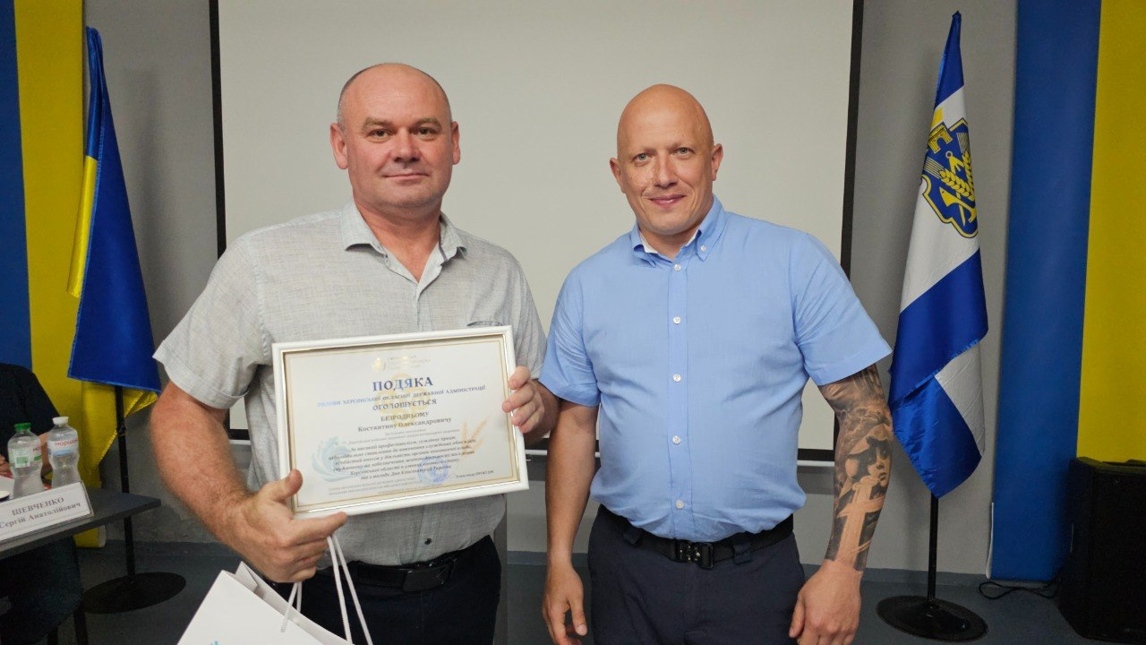 Двоє чоловіків стоять поруч у приміщенні з сірими стінами та тримають офіційну подяку в рамці, при цьому чоловік ліворуч вбраний у світлу сорочку з коротким рукавом. На задньому плані видно великий білий екран, державний прапор України та прапор Херсонської області.