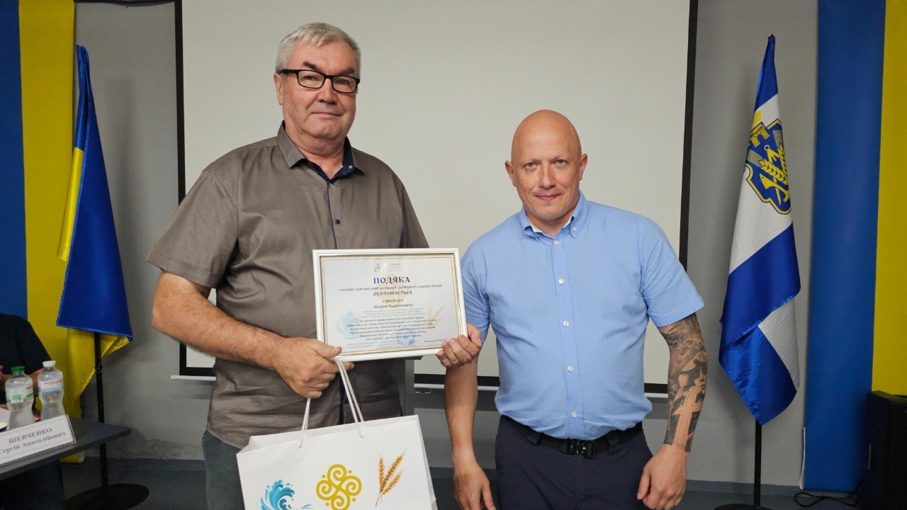 Двоє чоловіків стоять поруч у приміщенні з сірими стінами та тримають офіційну подяку в рамці, при цьому чоловік ліворуч в окулярах та сірій сорочці з коротким рукавом. На задньому плані видно великий білий екран, державний прапор України та прапор Херсонської області.
