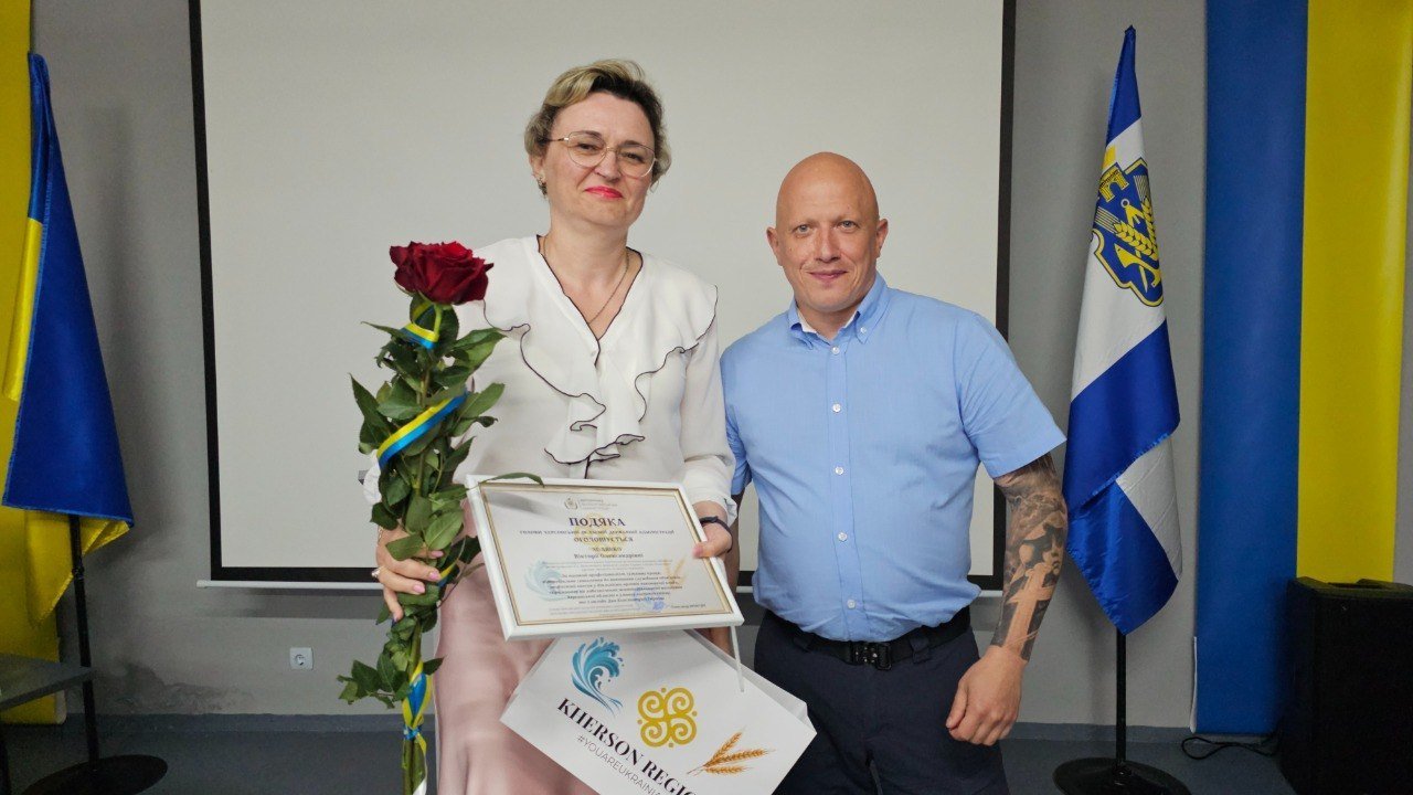 Чоловік та жінка в окулярах і світлій блузі стоять поруч у приміщенні з сірими стінами та тримають офіційну подяку в рамці. Жінка також тримає букет червоних троянд і фірмовий паперовий пакет, а на задньому плані видно великий білий екран, державний прапор України та прапор Херсонської області.