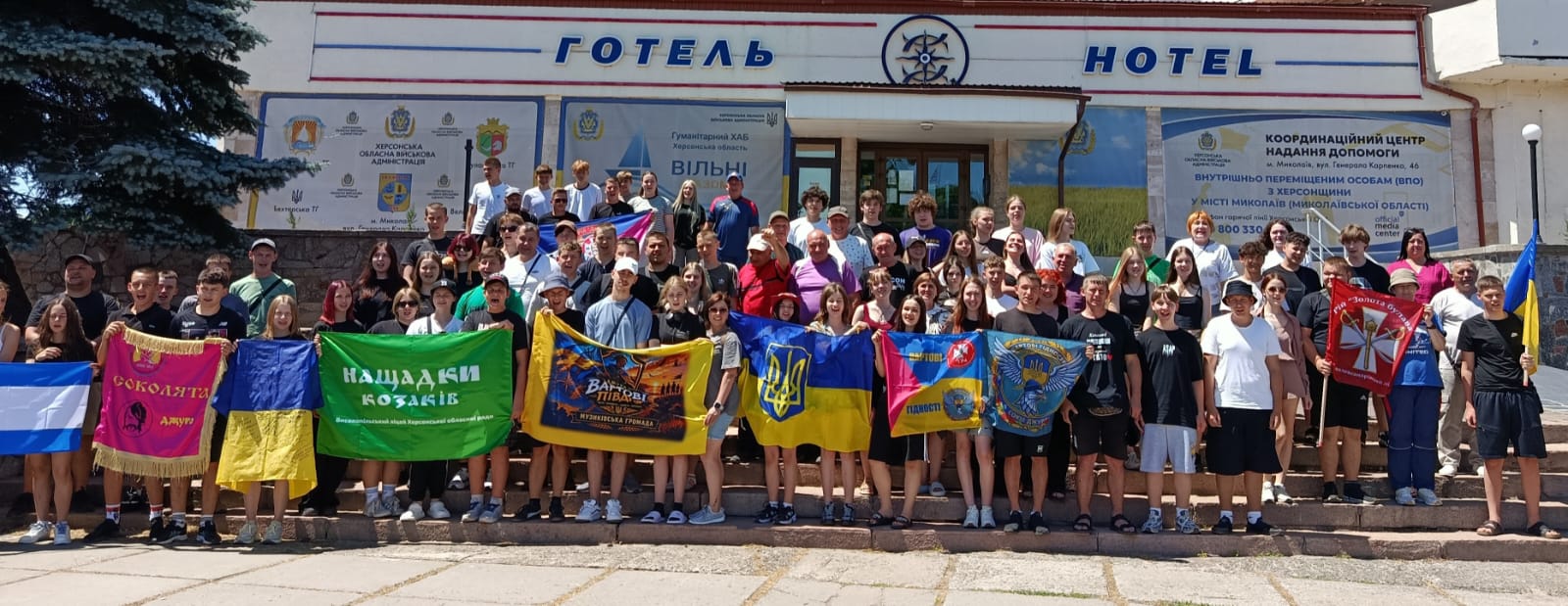 Велика група людей різного віку стоїть на сходах перед будівлею з написами «ГОТЕЛЬ» та «HOTEL». Вони тримають у руках численні прапори, серед яких державні прапори України, прапори з гербом та різнокольорові стяги з написами та символікою громад.