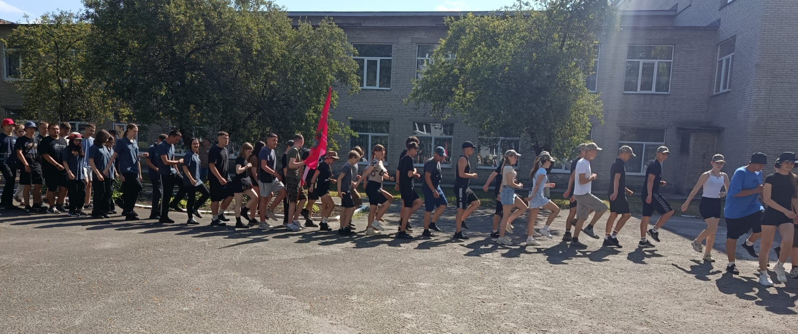 Велика група молоді у повсякденному та спортивному одязі марширує або крокує шеренгою по асфальтованому подвір'ю на тлі двоповерхової цегляної будівлі та зелених дерев. Один із учасників попереду несе великий яскраво-рожевий прапор.