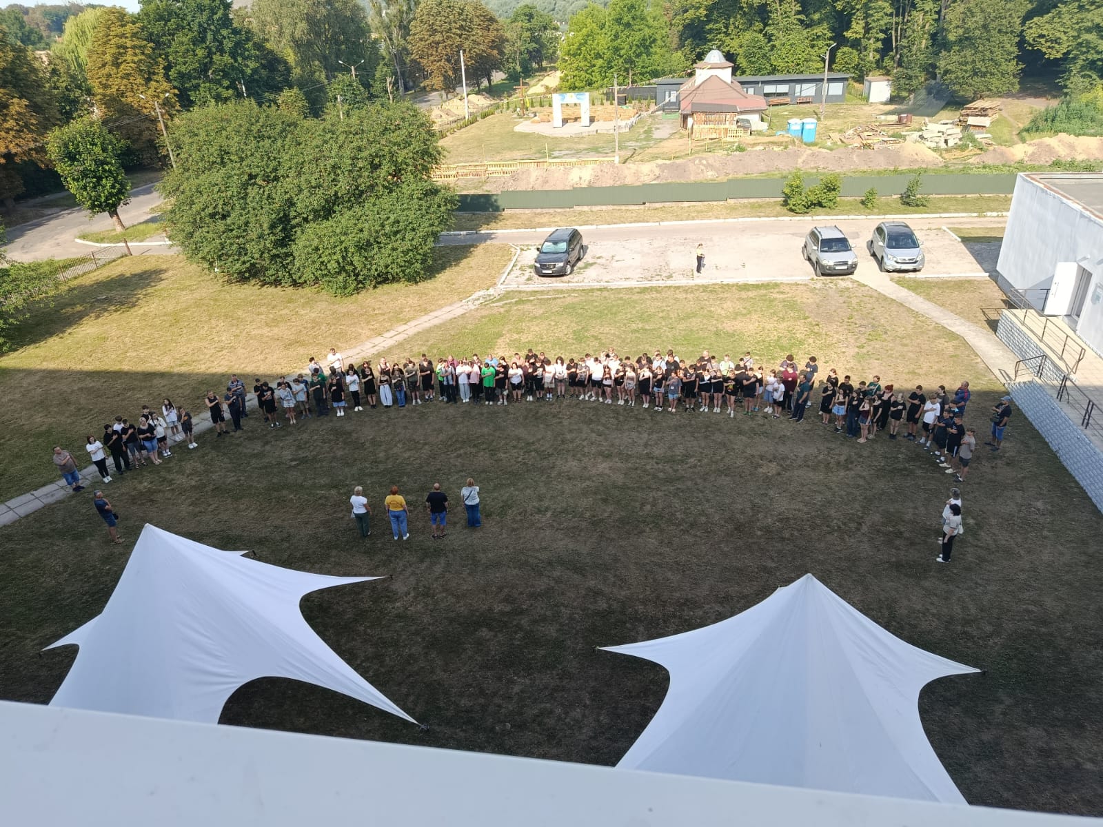 Велика група людей вишикувалася півколом на зеленому газоні, вид зверху. На передньому плані видно два великі білі намети-зірки, а на задньому плані — парковку з автомобілями, дерева та будівельний майданчик.