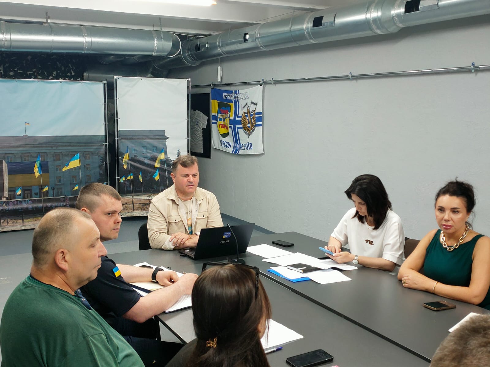 Нарада за довгим робочим столом в офіційному приміщенні, де присутні чоловіки та жінки у повсякденному одязі та формених сорочках з українською символікою.