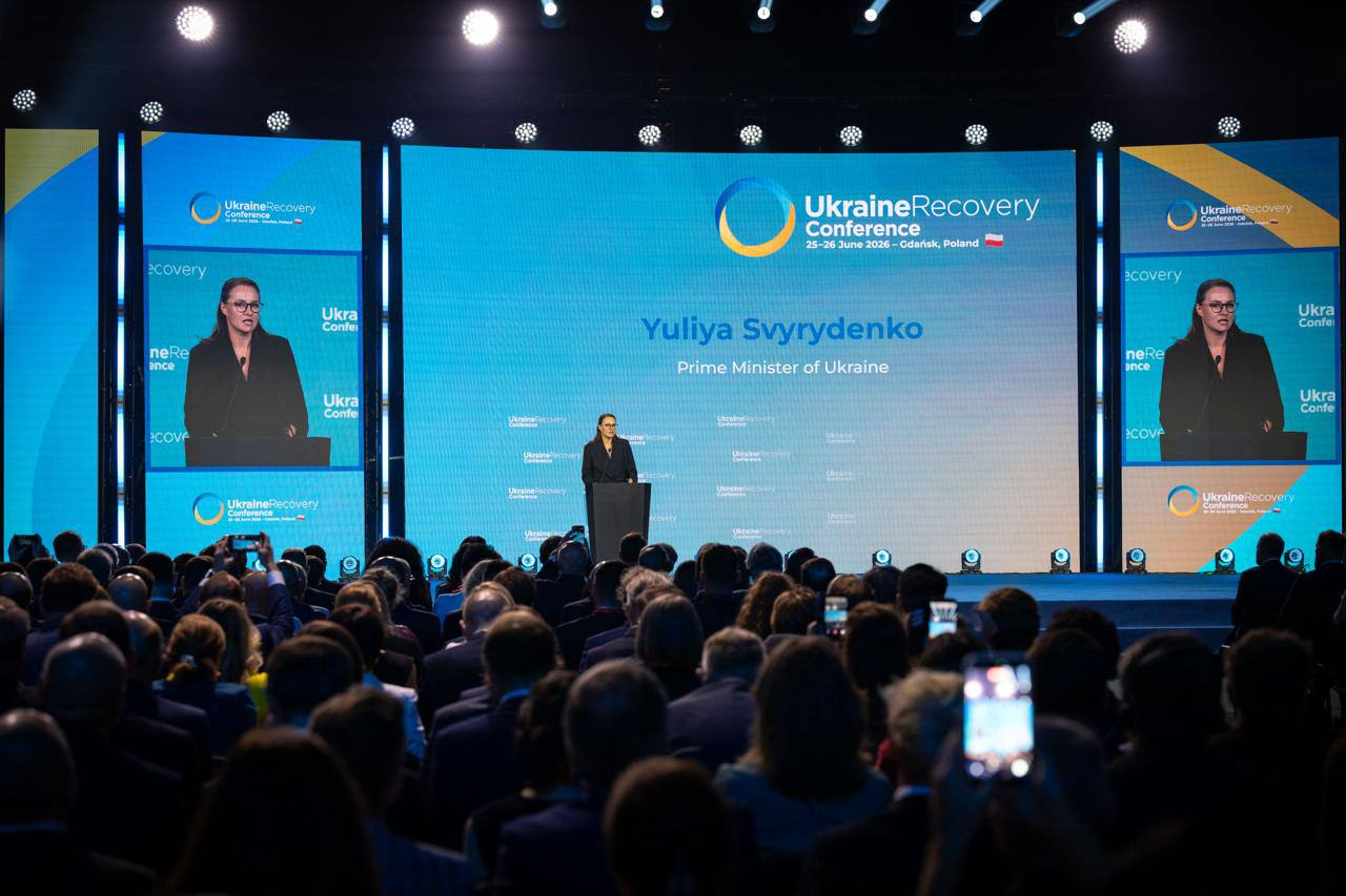 Виступ жінки за трибуною на масштабній офіційній конференції перед великою аудиторією в залі. На великому синьому екрані позаду неї розміщено написи англійською мовою та логотипи заходу.