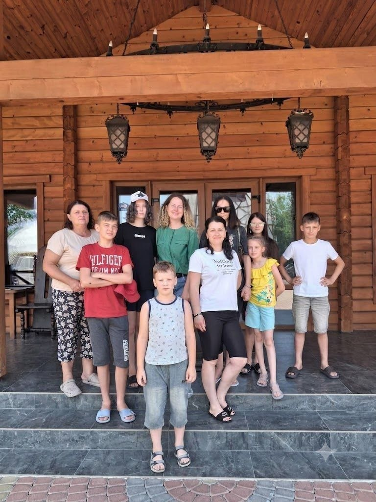 Група дітей та дорослих у літньому повсякденному одязі позує для спільного фото на ганку великого дерев'яного будинку зі зрубу.