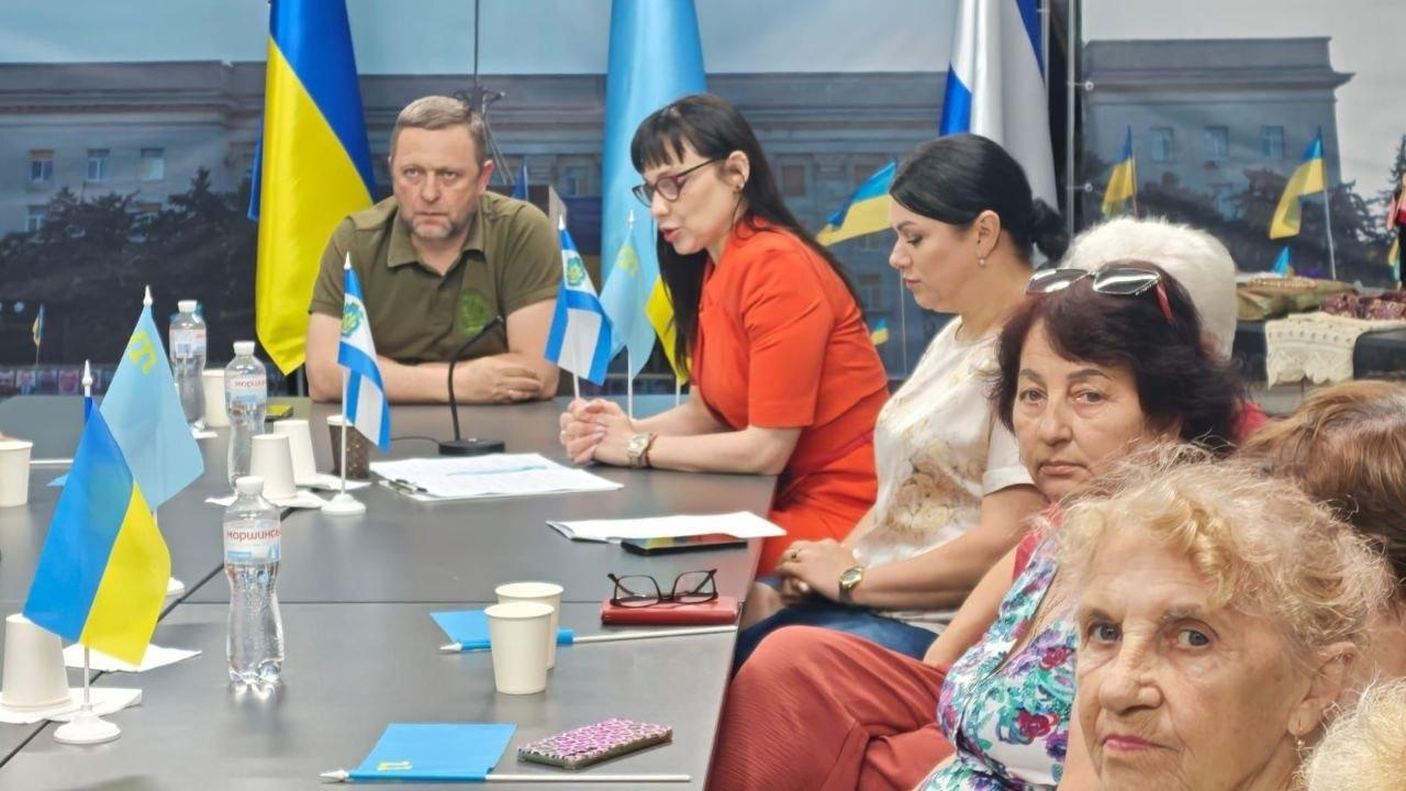 Офіційна зустріч у приміщенні, де жінка в яскраво-помаранчевій сукні виступає перед присутніми за довгим столом на тлі прапорів України та Херсона.