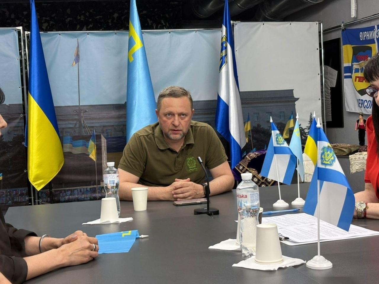 Чоловік у футболці кольору хакі сидить на чолі великого столу під час офіційної зустрічі, де перед ним встановлені настільні прапорці України та міста Херсона.