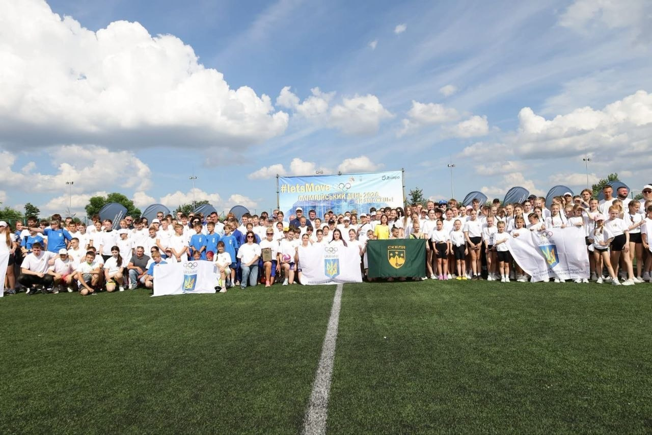 Велика група дітей та молоді у спортивній формі позує для спільного фото на зеленому штучному полі під час заходу «Олімпійський день 2026», тримаючи прапори з олімпійською символікою.