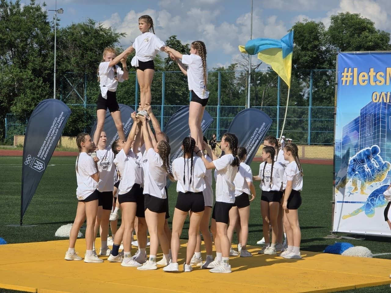 Група дівчат-черлідерок у білих футболках та чорних шортах будує акробатичну піраміду на жовтому маті просто неба під час спортивного заходу.