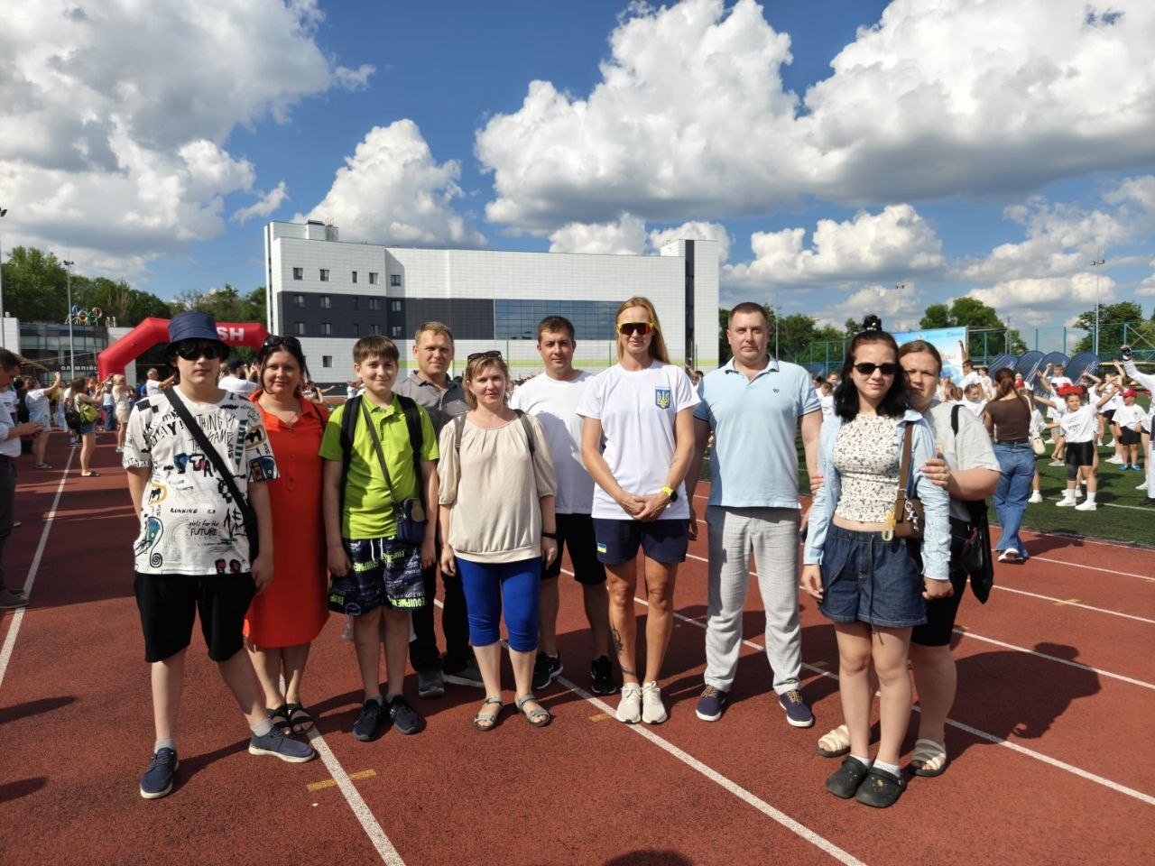 Група людей у літньому повсякденному та спортивному одязі позує для спільного фото на біговій доріжці відкритого стадіону на тлі сучасної будівлі спортивного комплексу.
