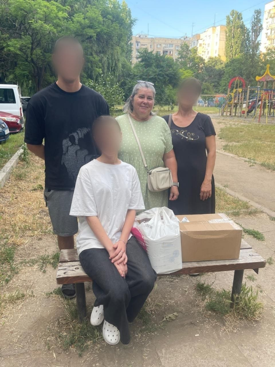 Група людей позує для фото на вулиці біля дерев'яної лави з картонною коробкою та пакунком гуманітарної допомоги; обличчя трьох осіб на світлині штучно розмиті.