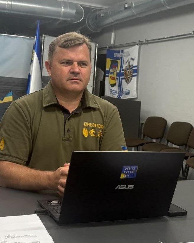Чоловік у зеленій тенісці з написом «Kherson Region» сидить за столом перед відкритим ноутбуком ASUS у робочому кабінеті чи офісі.