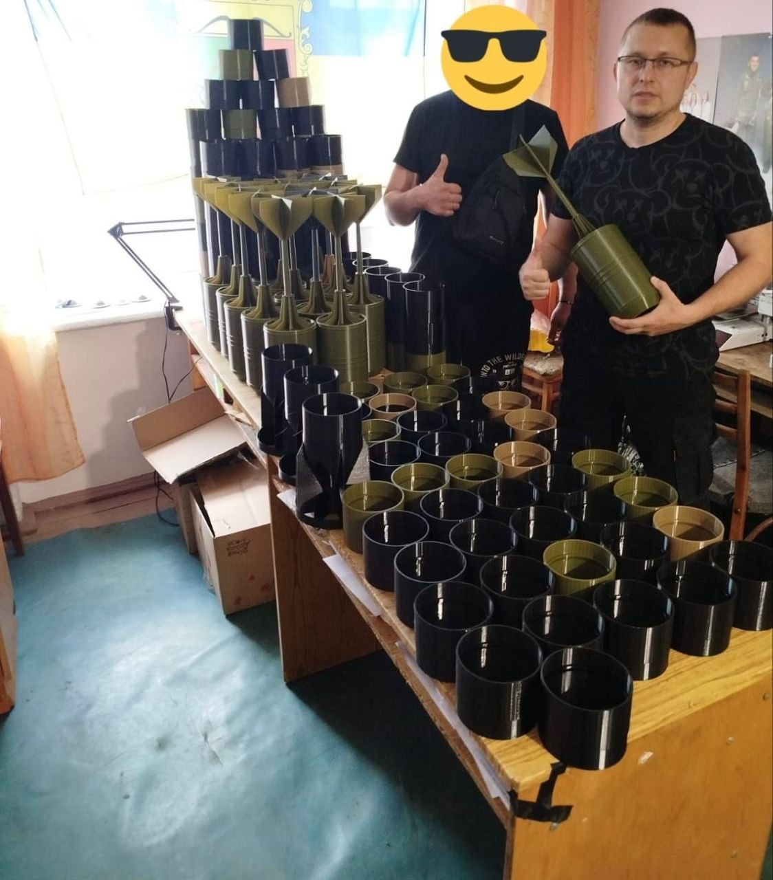 Двоє чоловіків стоять у приміщенні біля столу, щільно заставленого чорними та зеленими пластиковими елементами для боєприпасів. Обличчя одного з них приховане жовтим емодзі, а інший тримає в руках готовий виріб із хвостовим стабілізатором. На задньому плані навколо столу видно картонні коробки та швейну машинку.