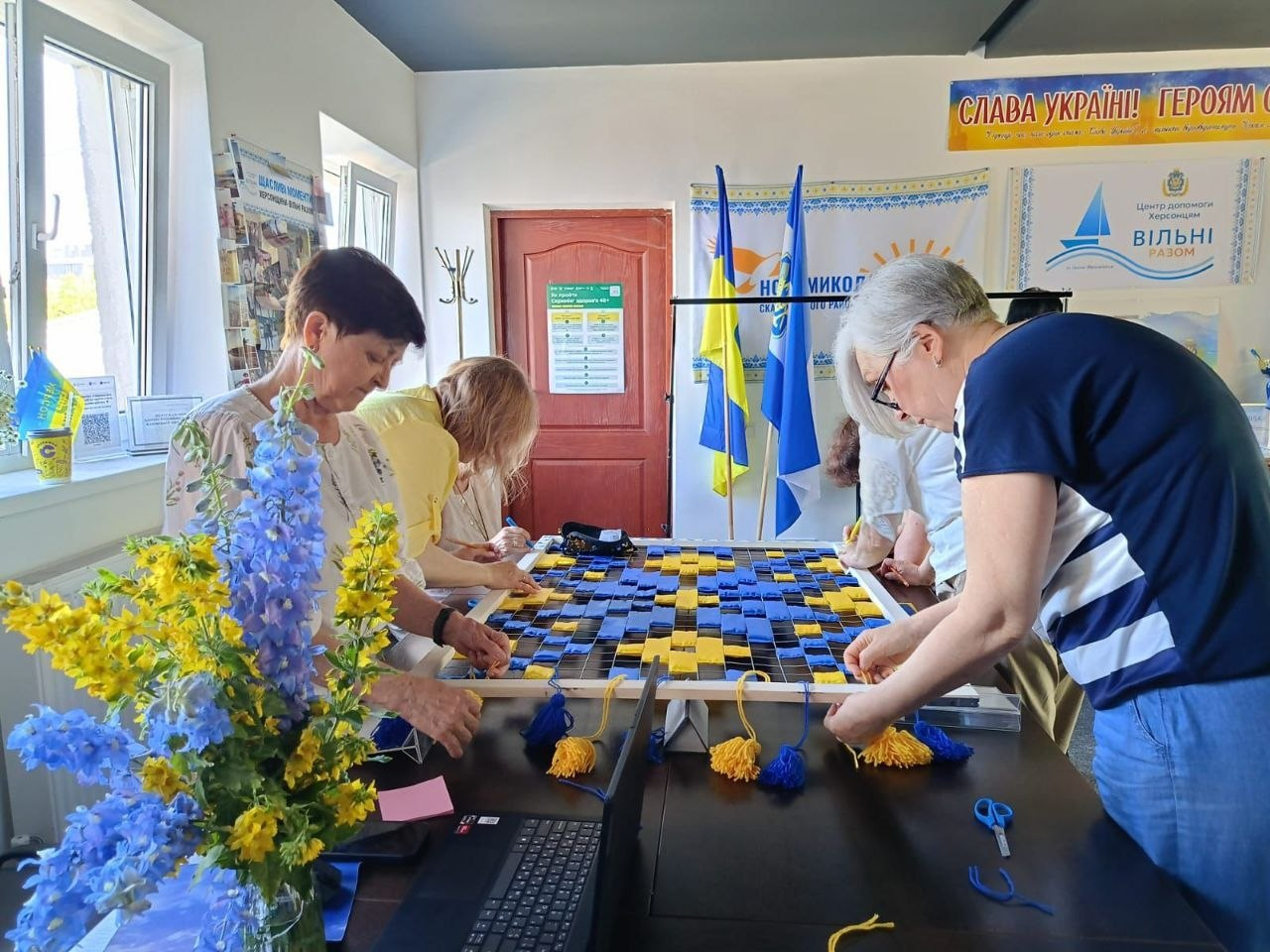 Група людей за столом у приміщенні спільно плете маскувальну сітку з жовтих та синіх стрічок. На передньому плані стоїть ваза з патріотичним букетом із жовтих та синіх квітів, а на стінах видніються прапори України та плакати з патріотичними гаслами.