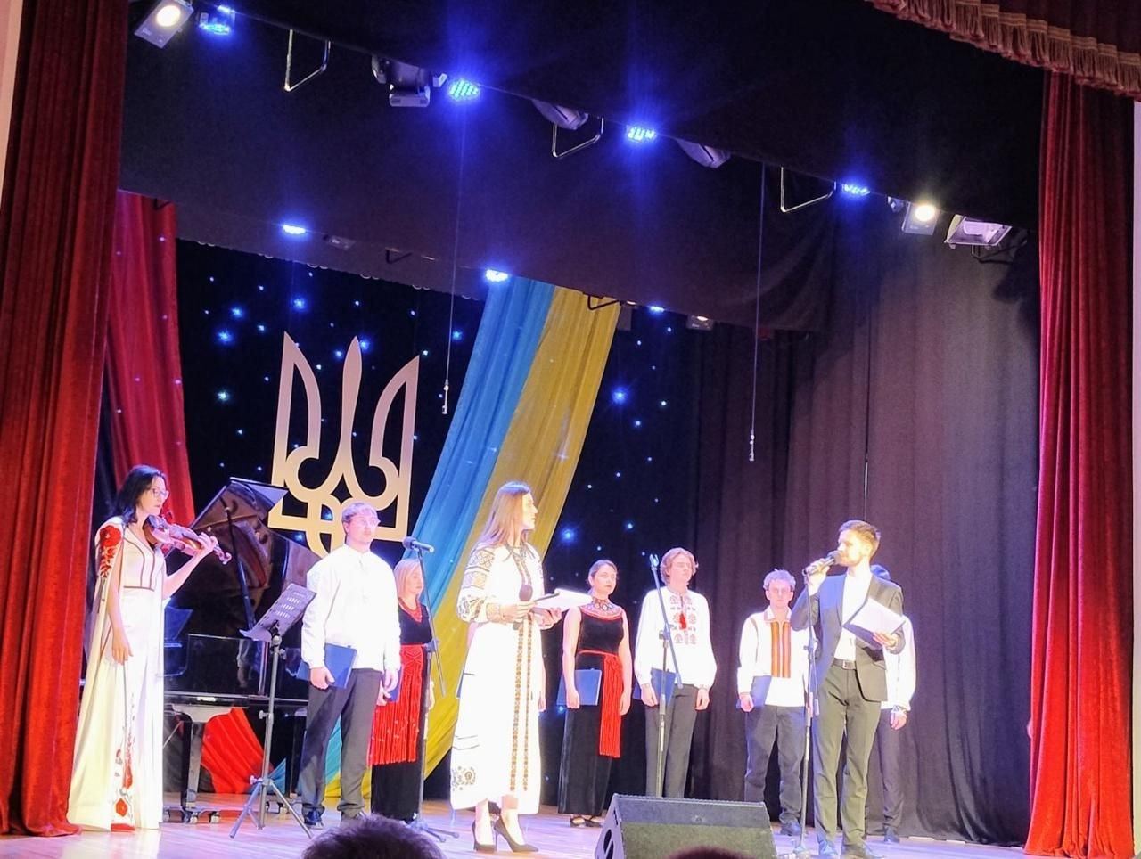 Група артистів виступає на освітленій сцені театру або будинку культури. На задньому плані сцени розміщено великий герб України (Тризуб), державний прапор та зоряне тло, а ліворуч стоїть рояль із скрипалькою у вишитій сукні.