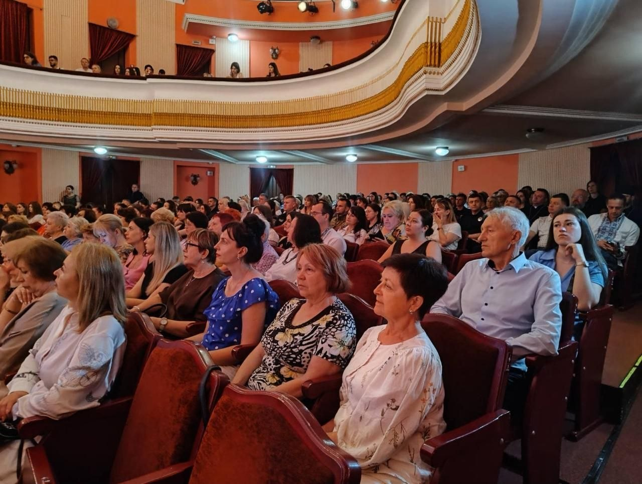 Глядацька зала театру або будинку культури, заповнена людьми, які сидять у кріслах і дивляться в бік сцени. Над партером видно ярус балкона з іншими глядачами, а інтер'єр виконано в помаранчевих, білих та коричневих тонах.